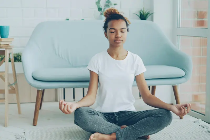 A woman sitting cross-legged on a rug at home, eyes closed, beside an armchair and house plants