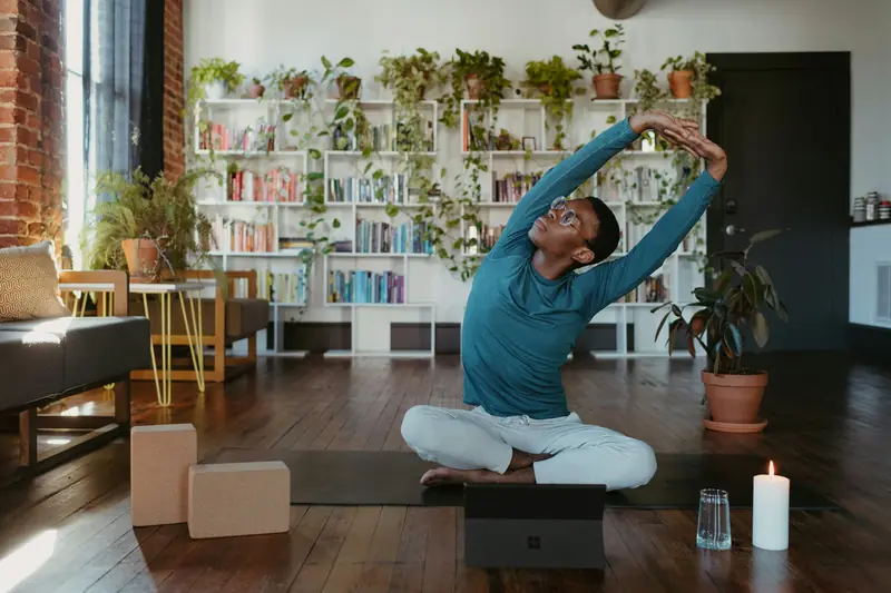 A man stretching sideways on a yoga mat in a plant-filled living room with yoga blocks and a candle nearby